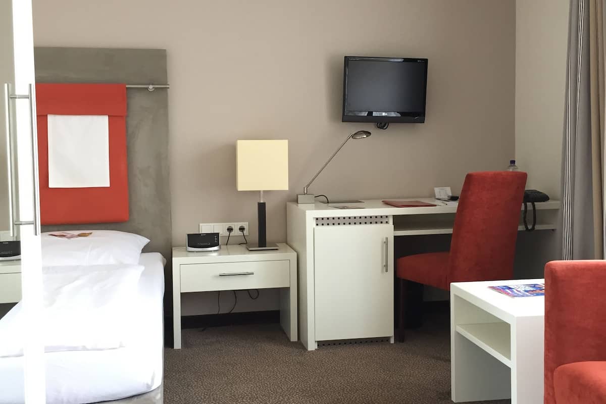 Habitación de hotel con cama, escritorio, silla, televisor y una pequeña mesa con lámpara.