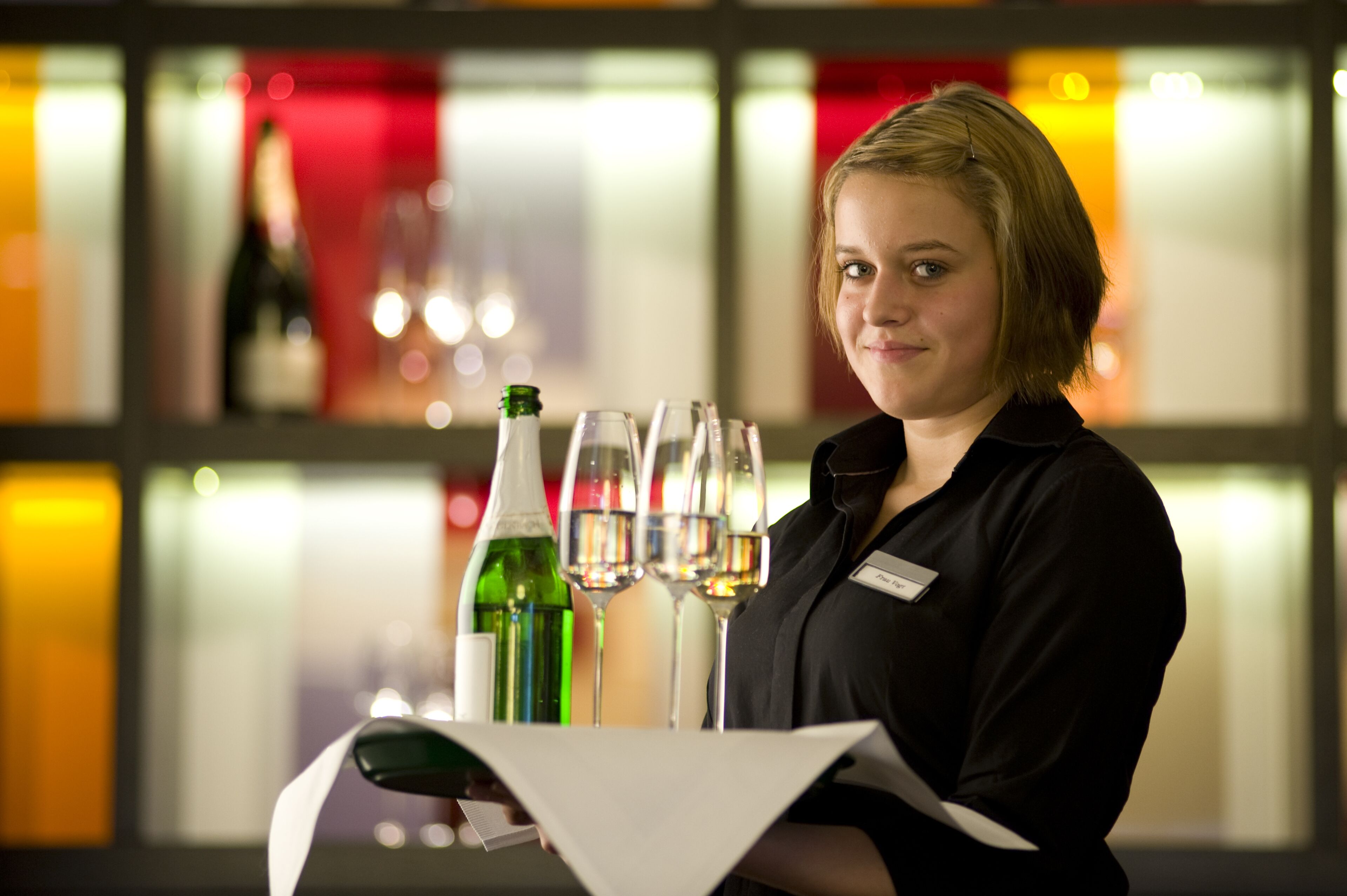 A person holding a tray with wine bottles and glasses, with a blurred background of a room with windows and lights.