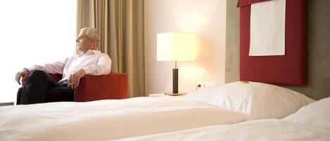 Una habitación de hotel con una cama, un sillón rojo, una lámpara y un ventanal con cortinas.