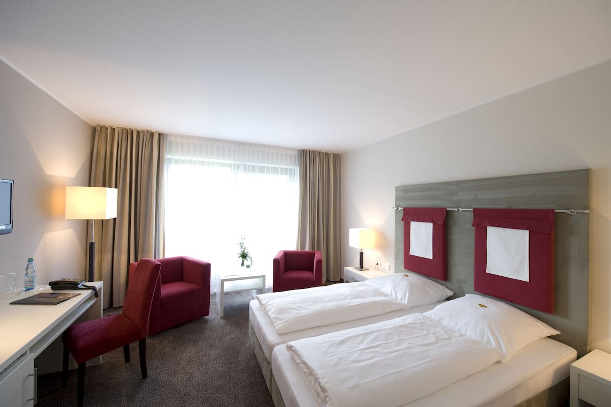Habitación de hotel con dos camas, un escritorio, una silla, una lámpara y una ventana con cortinas.