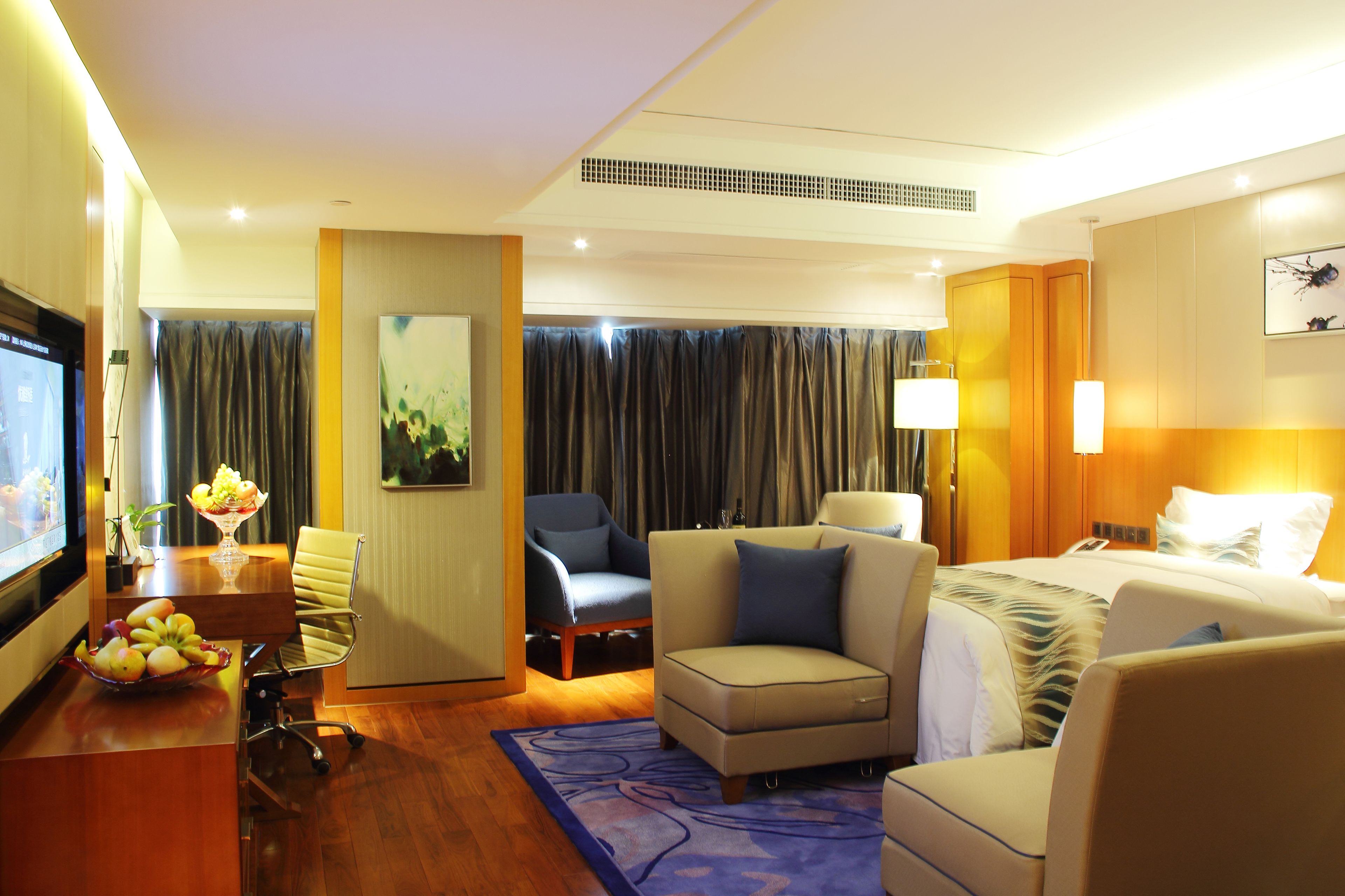 A hotel room with a bed, two armchairs, a desk with a fruit bowl, a TV, and a large window with curtains.