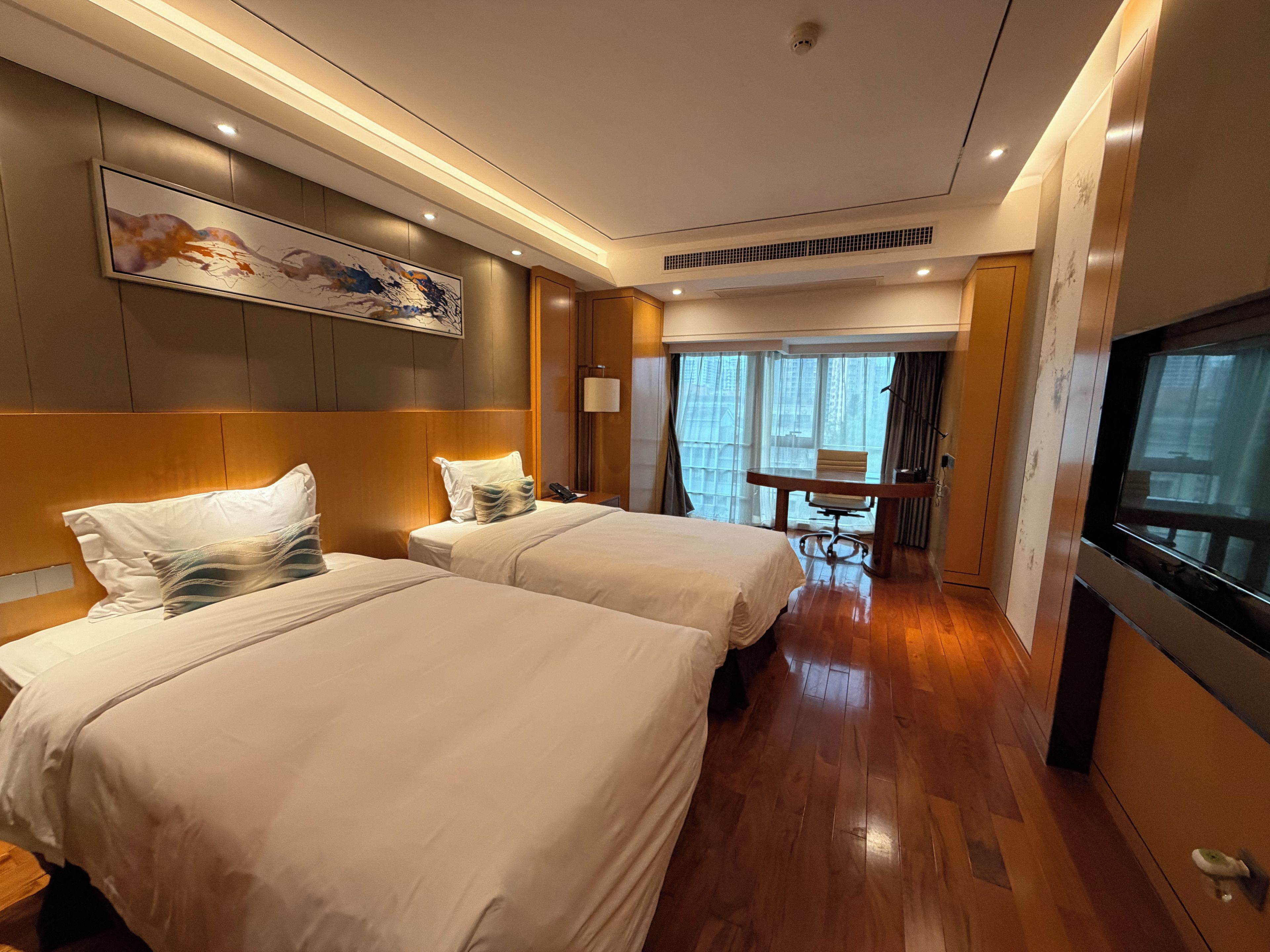 A hotel room with two beds, a desk, a chair, a TV, and a painting on the wall.