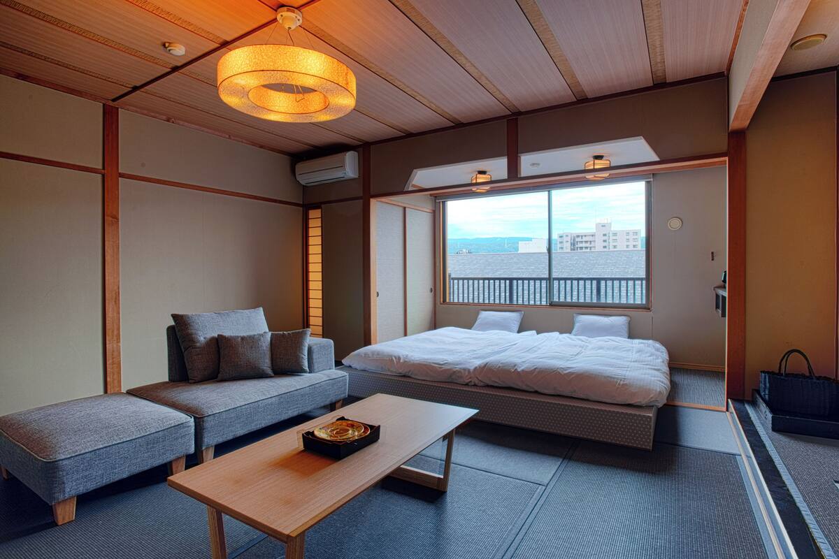 Une pièce comprenant un lit, un canapé et une table, avec vue sur les bâtiments par une fenêtre.