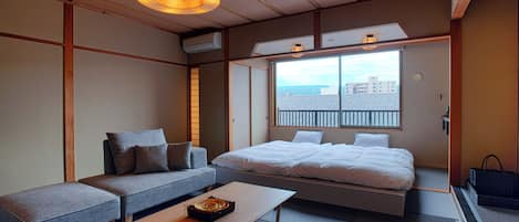 A room with a bed, sofa, and table, with a view of buildings through a window.