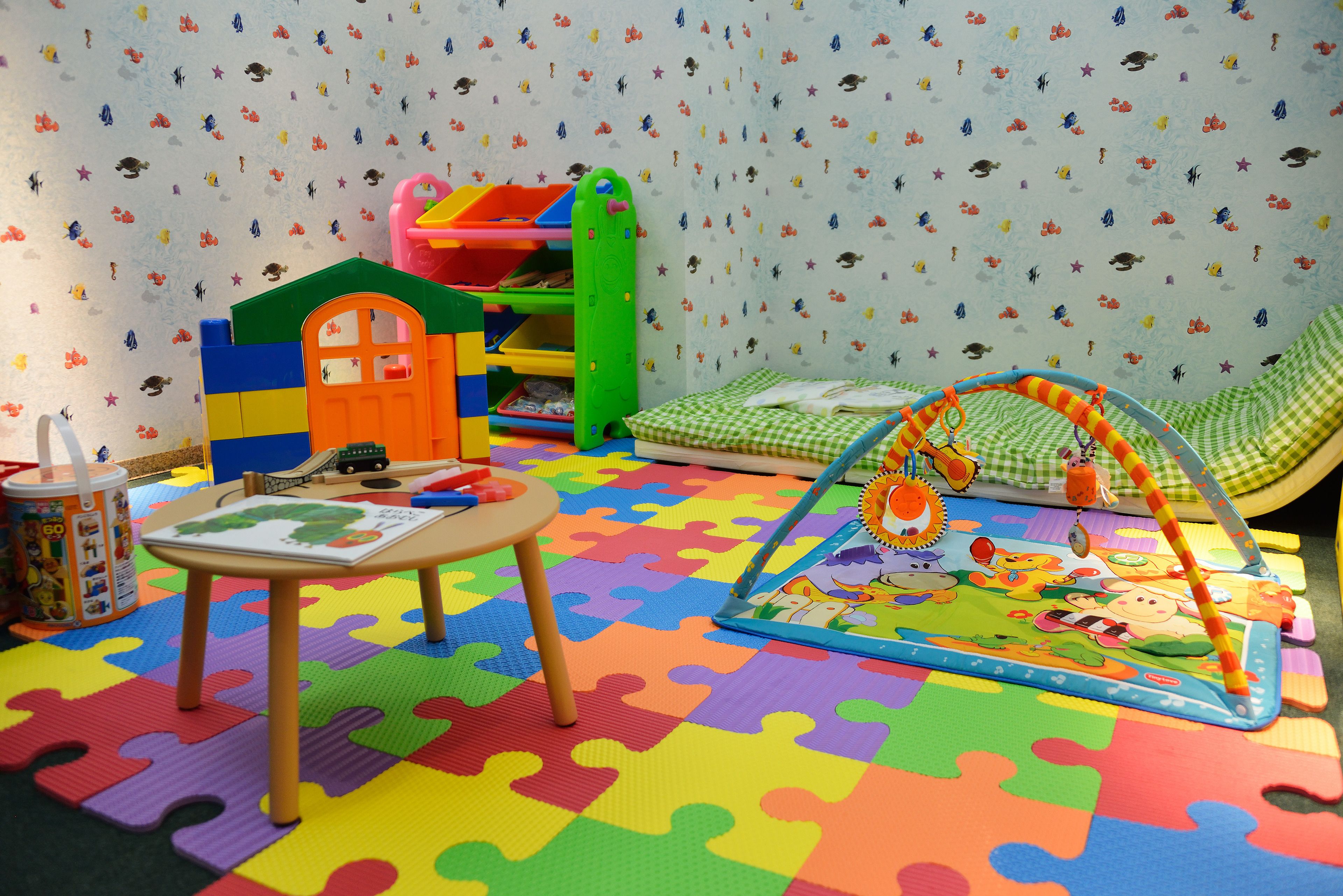 Une salle de jeux pour enfants avec un tapis de jeu coloré, une petite table, une maisonnette et un jouet suspendu.