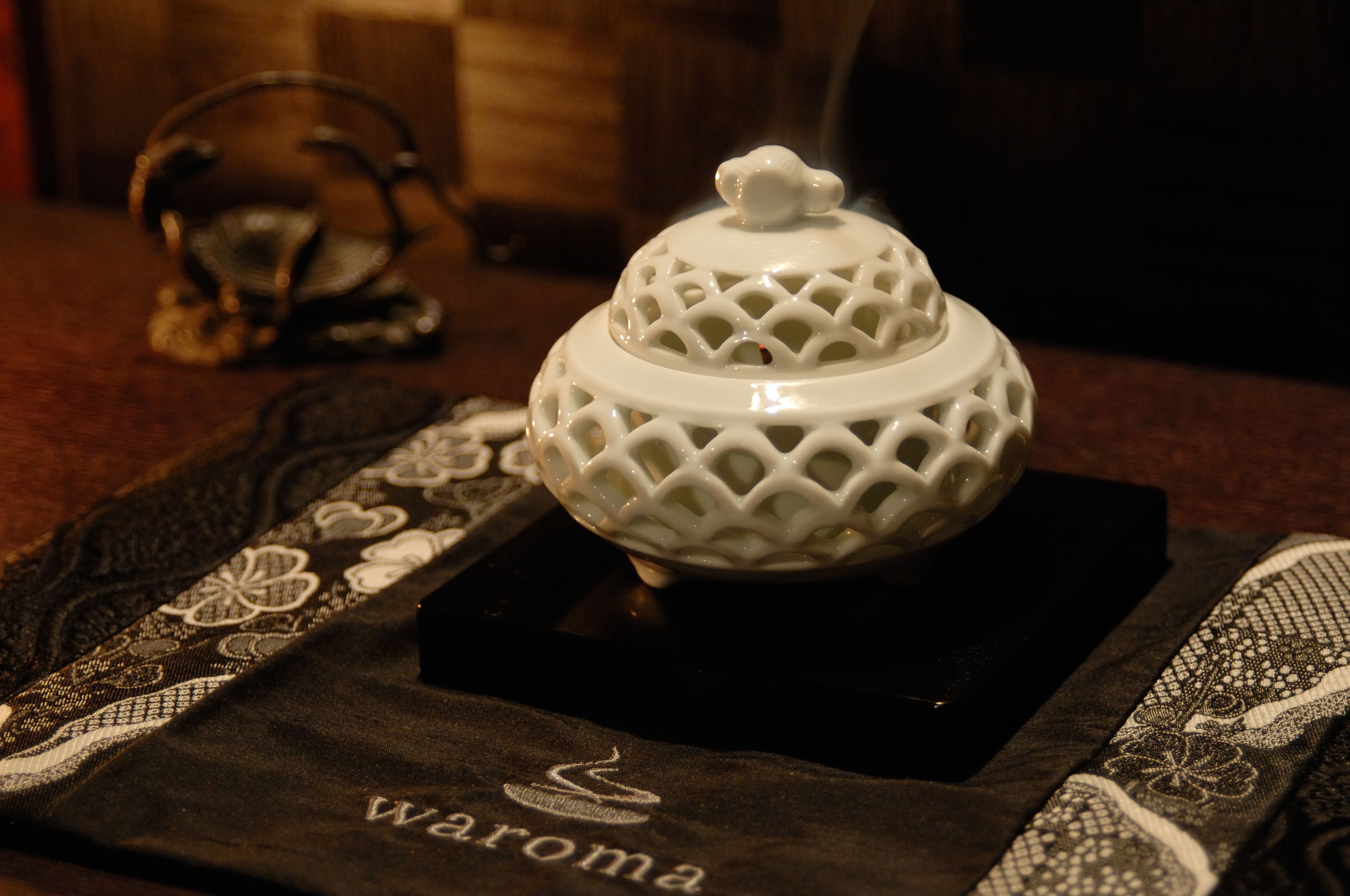 A white ceramic jar with a lid on a dark cloth with 'Waroma' embroidered on it.