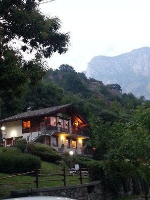 A chalet-style building with multiple balconies, surrounded by lush greenery and mountains in the background.