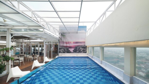 An indoor swimming pool with checkered tiles, surrounded by white lounge chairs and a city view through large windows. - Grand Tebu Hotel (Bandung)