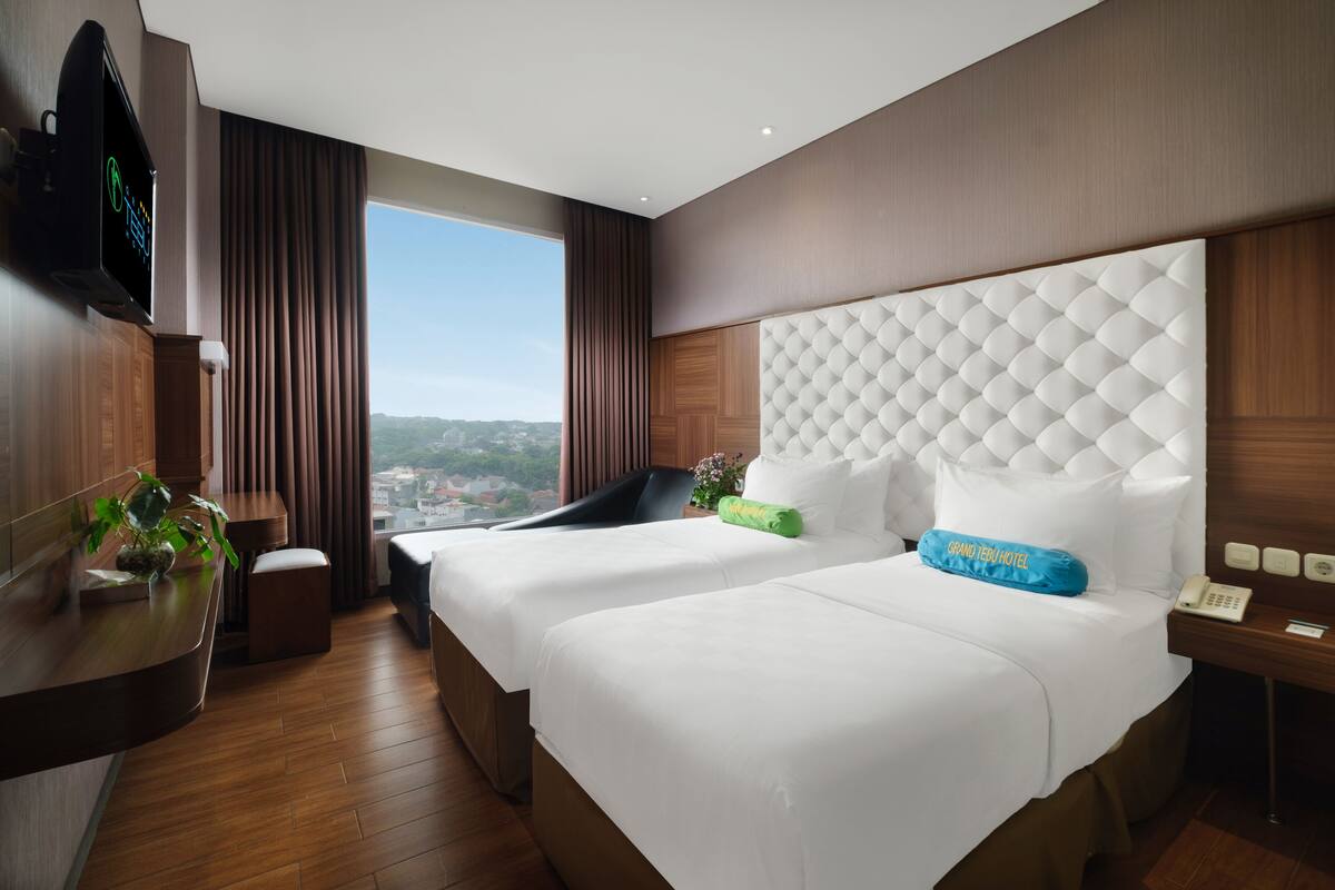 A hotel room with two beds, a TV, a desk, and a view of the city.