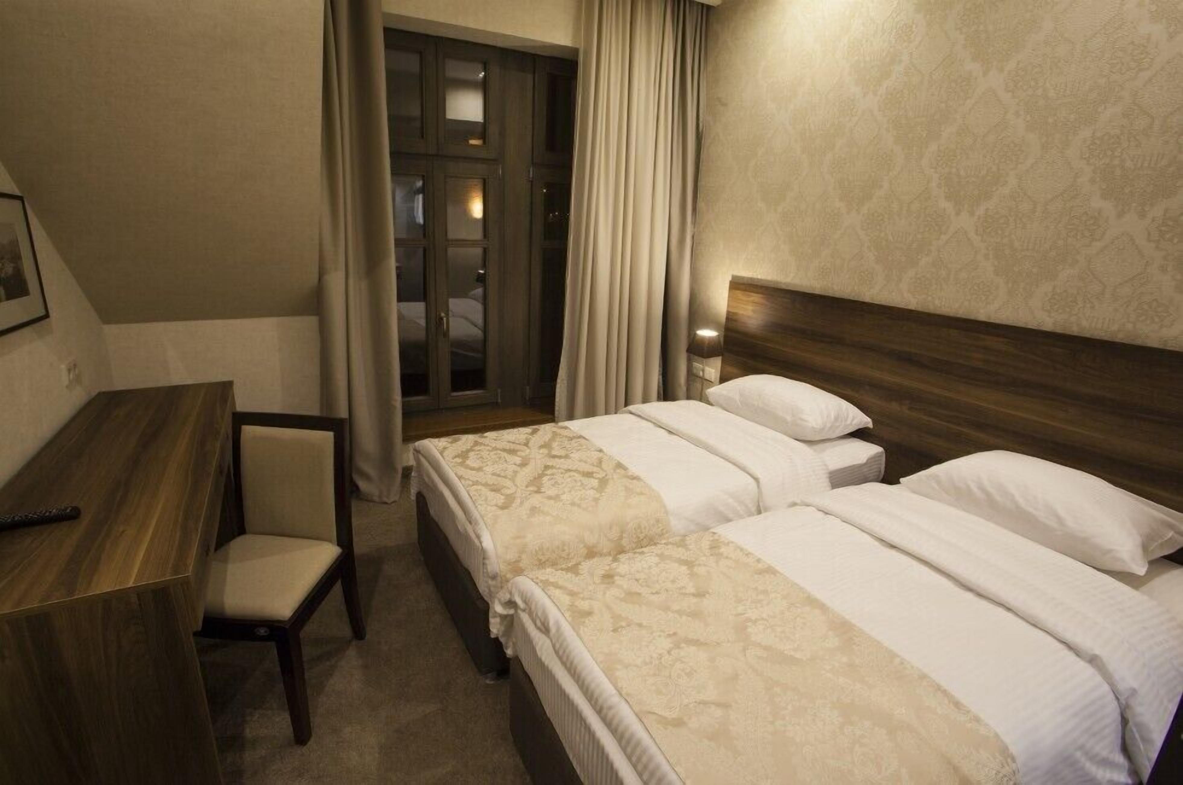 Une chambre d’hôtel avec un bureau en bois, deux lits avec du linge de lit blanc, une chaise et une fenêtre avec des rideaux.