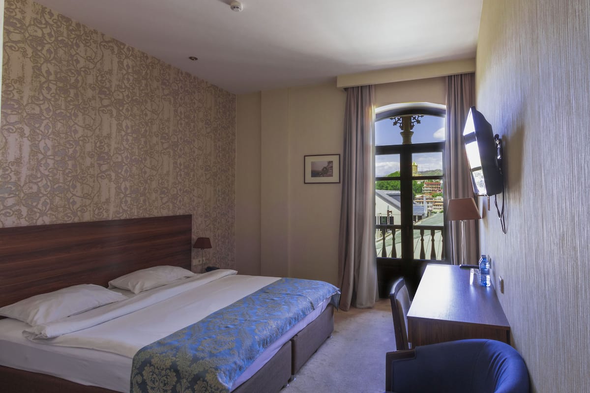 A hotel room with a bed, a desk, a chair, a TV, and a balcony with a view of buildings.