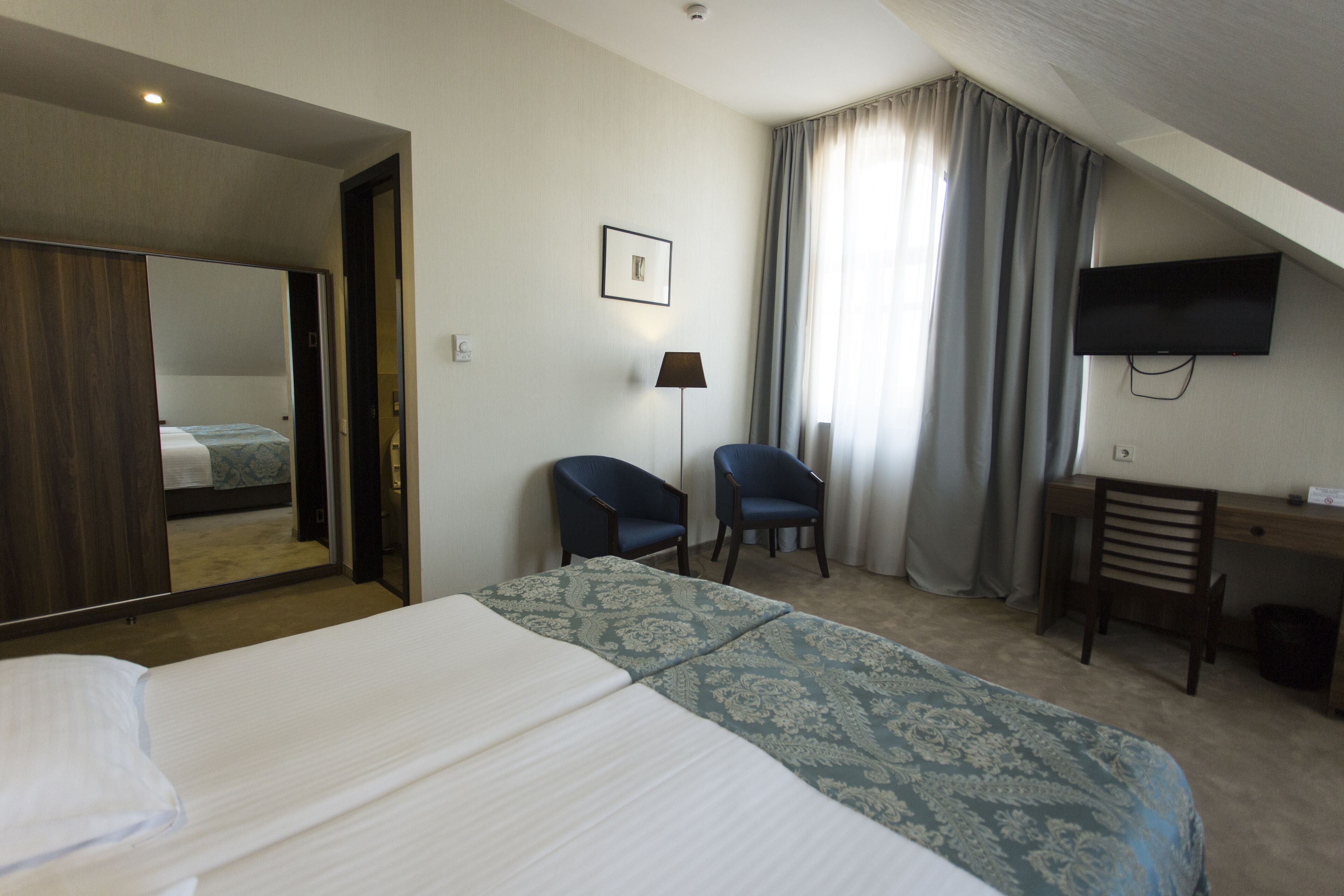 A hotel room with a bed, two chairs, a desk, a television, and a large mirror.