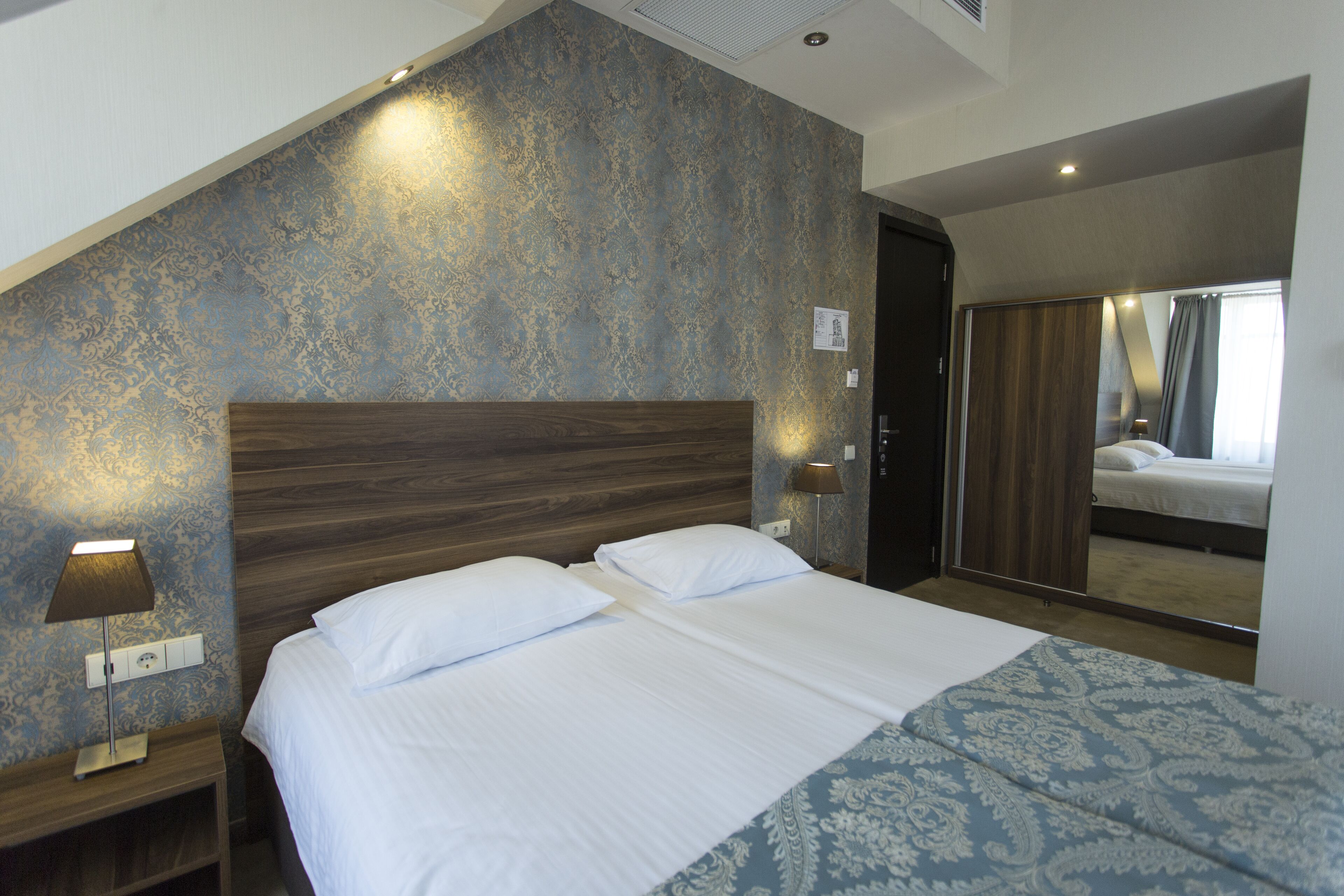 A hotel room with a large bed, a wooden headboard, a nightstand with a lamp, and a mirror on the wall.