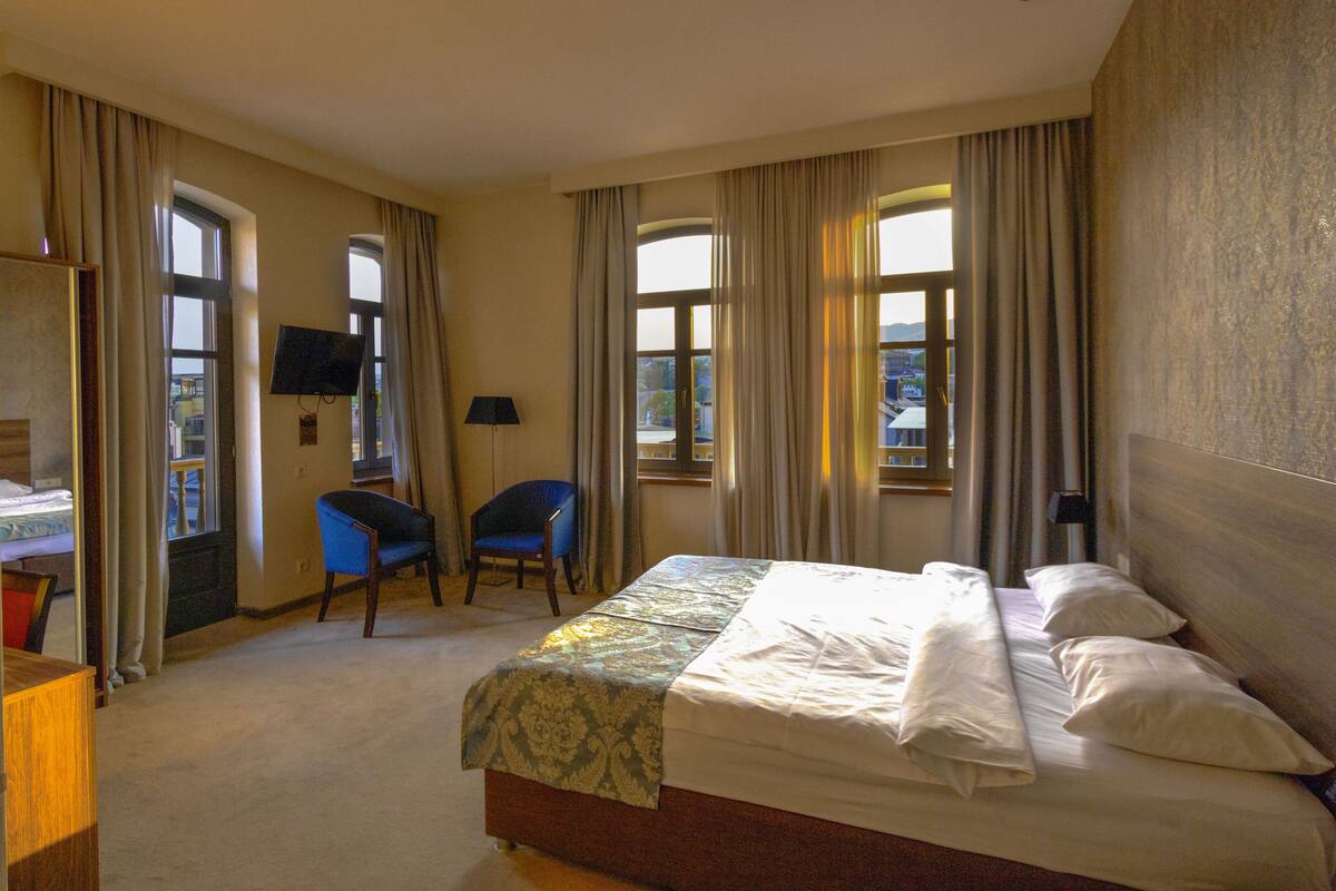 A hotel room with a bed, two chairs, a desk, a TV, and large windows.
