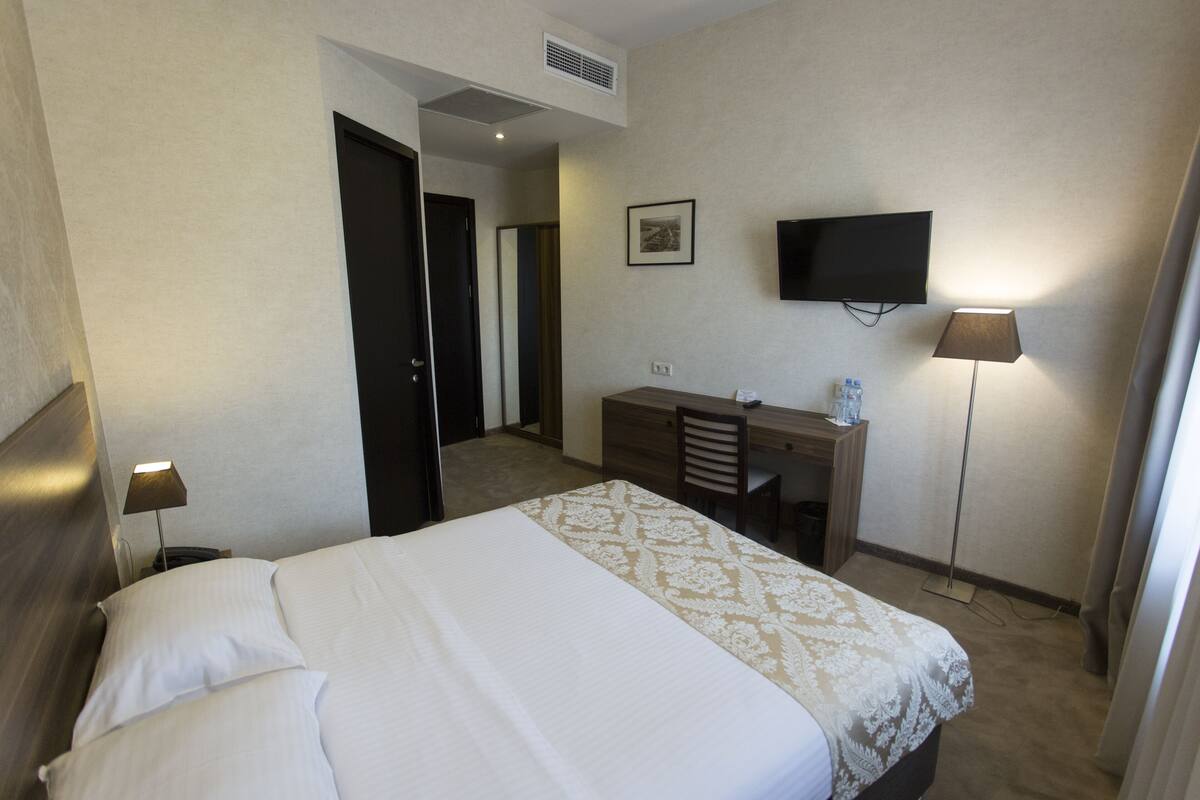 A hotel room with a bed, a desk, a chair, a TV, and a lamp.