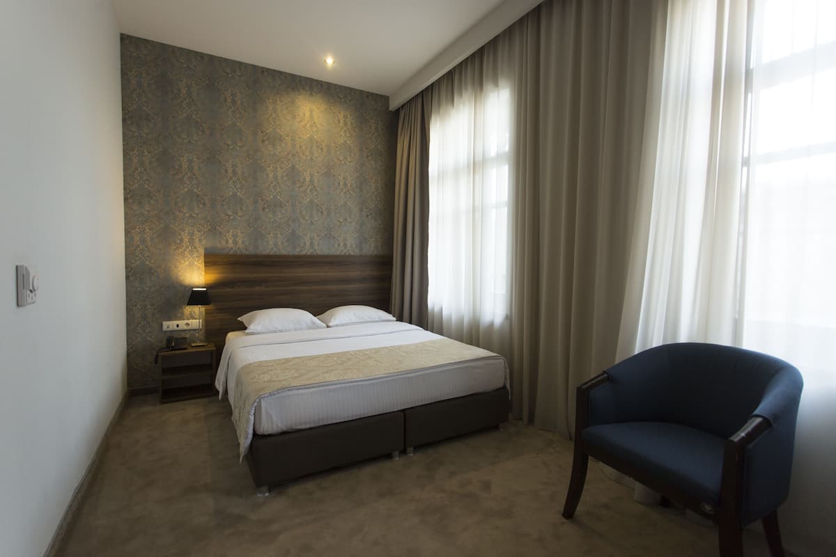 A hotel room with a bed, a chair, a nightstand with a lamp, and a window with curtains.