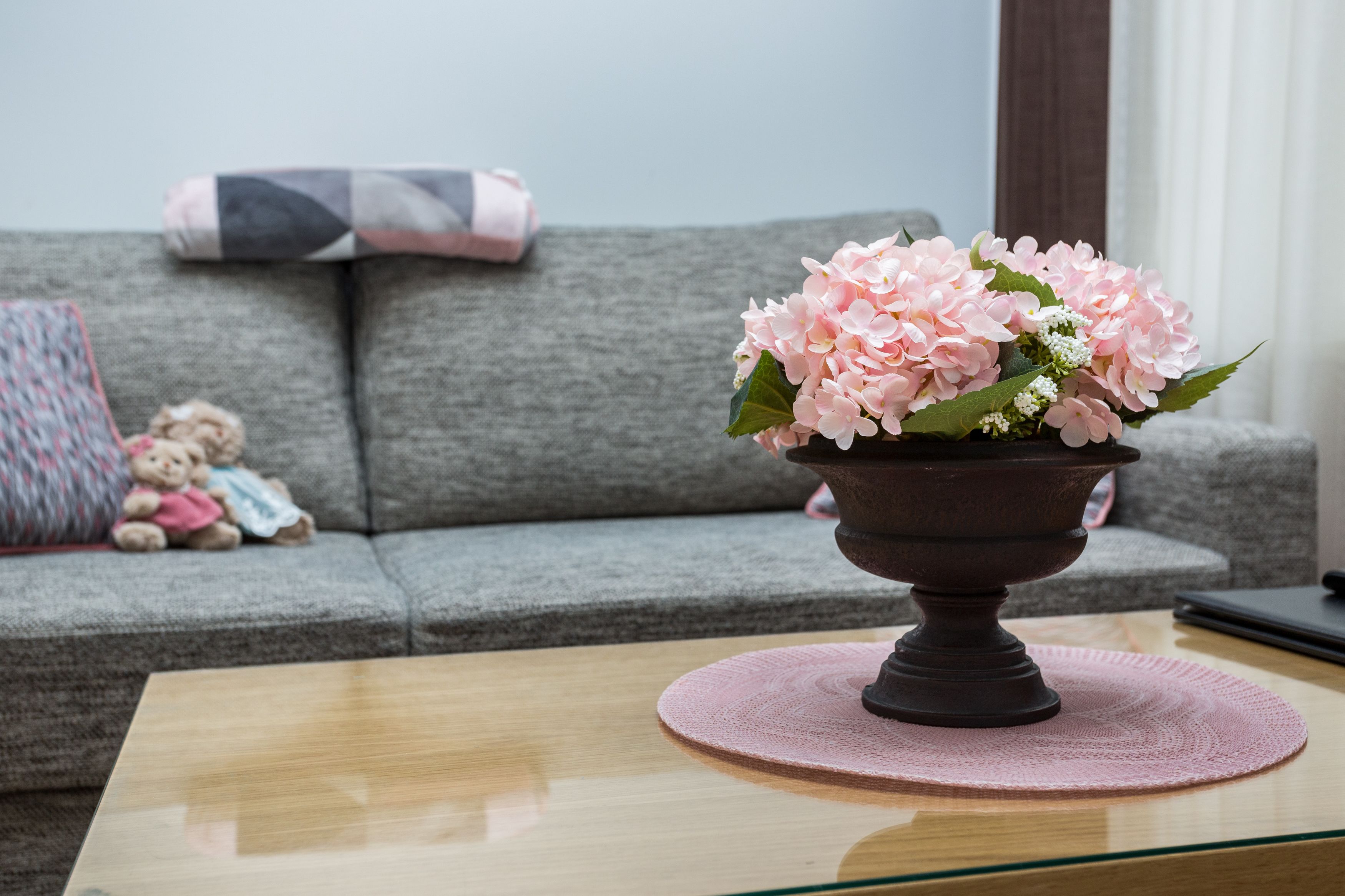 A living room with a grey sofa, a small table with a pink placemat, and a vase of pink flowers.