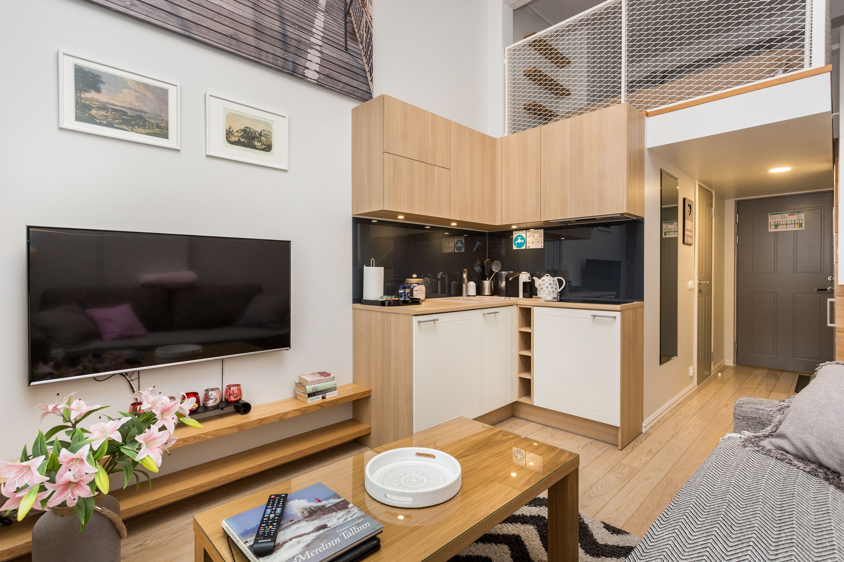 A modern living room with a flat-screen TV, wooden cabinets, a kitchen area, and a staircase leading to an upper level.