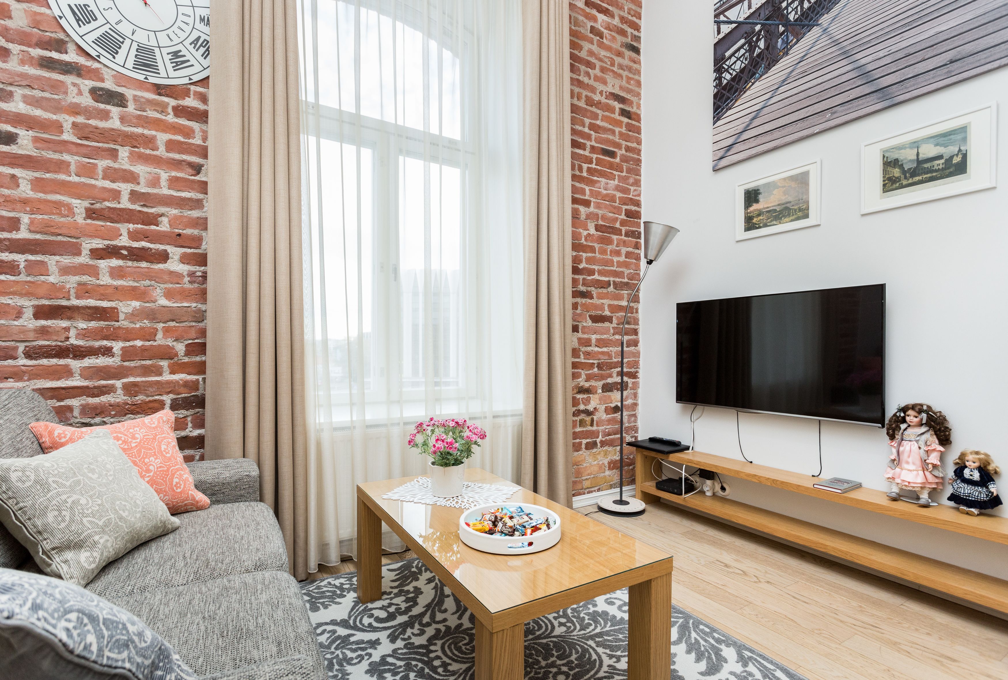 A living room with a brick wall, a large window with curtains, a flat-screen TV, a wooden coffee table, a sofa with patterned cushions, and a small shelf with decorative items.