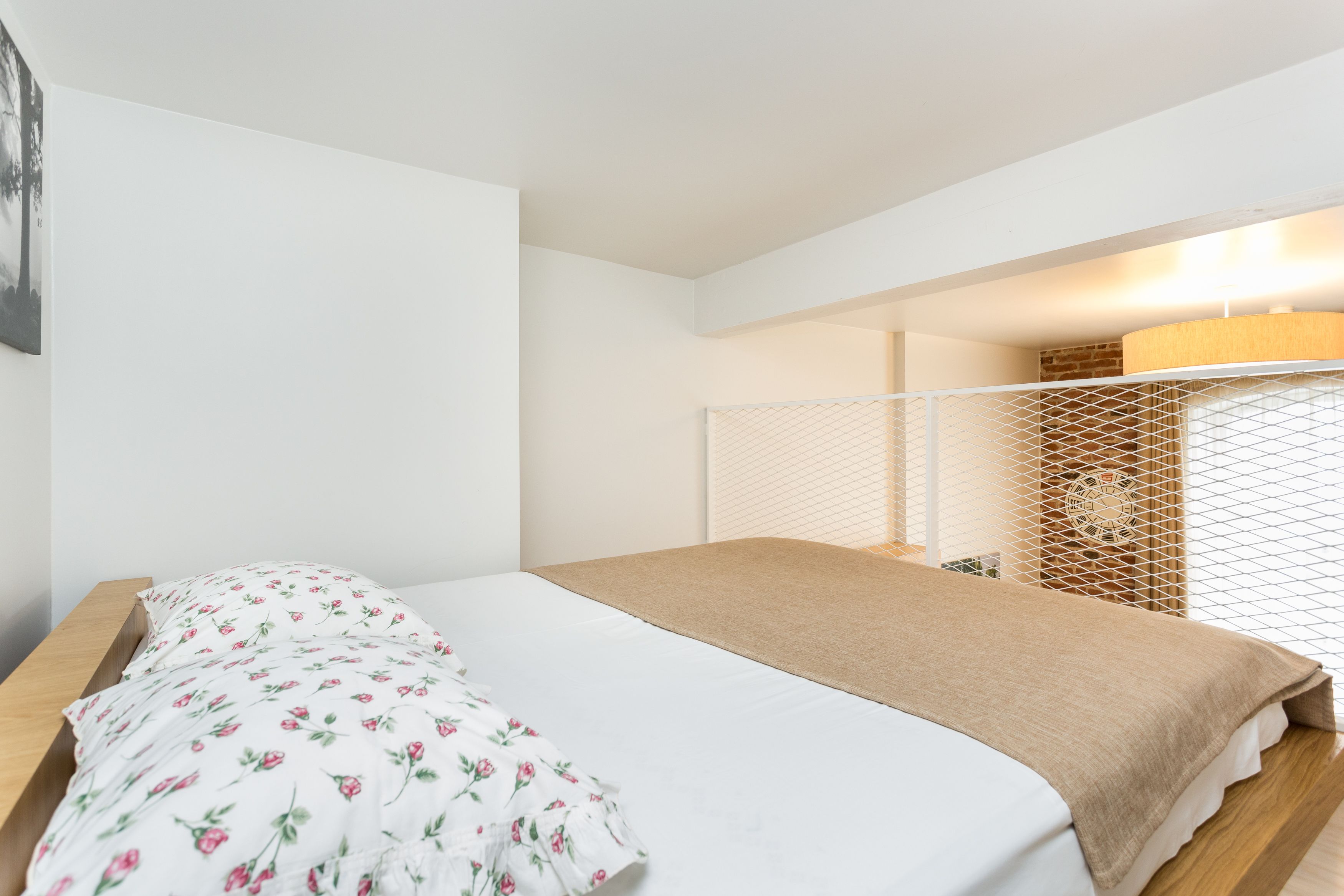 A bedroom with a bed, a wooden nightstand, a white wall, a window with blinds, and a lamp.