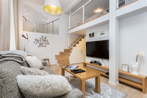 A modern living room with a sofa, coffee table, and a TV. There is a staircase with a railing, and a chandelier. - Angleterre Apartments (Tallinn)