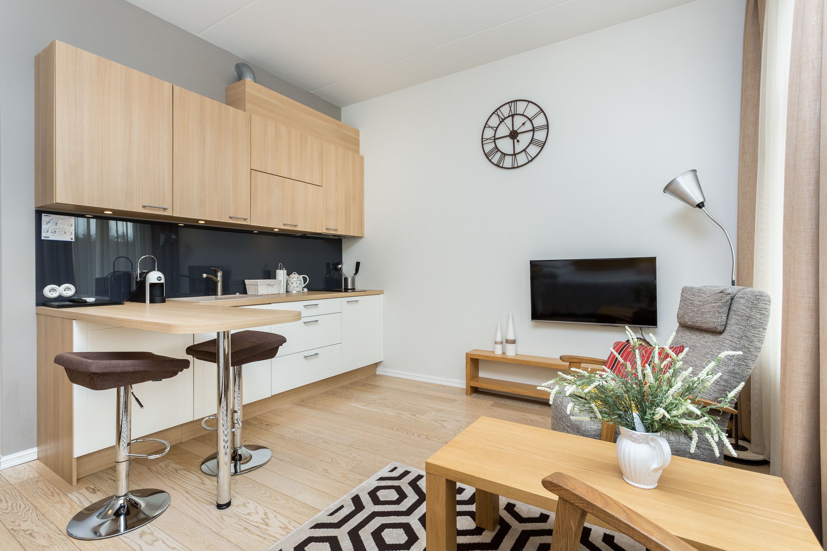 A modern kitchen with wooden cabinets, a bar stool, a dining table, and a TV in the living area.