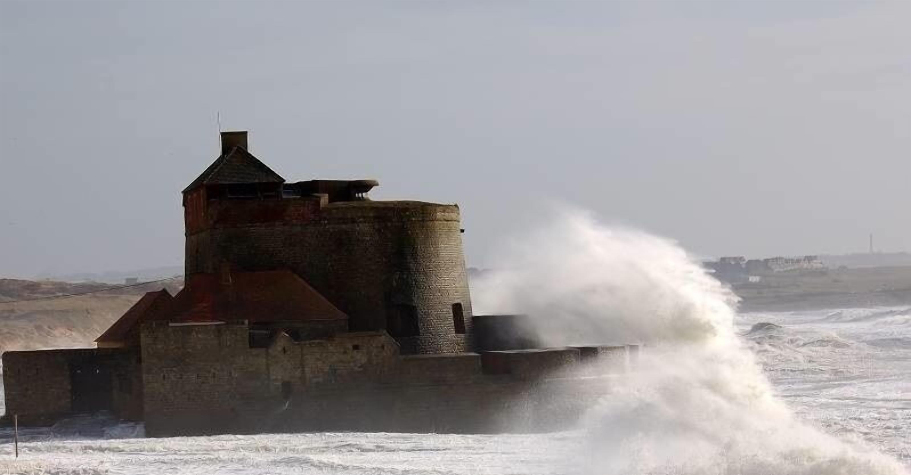 Eine Küstenbefestigung mit einem markanten Turm und einer großen Welle, die daran bricht.