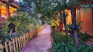 A narrow pathway flanked by lush greenery and traditional architecture, leading to a building with a red facade and ornate windows. - Gracious Bagan Hotel (Nyaung-U)