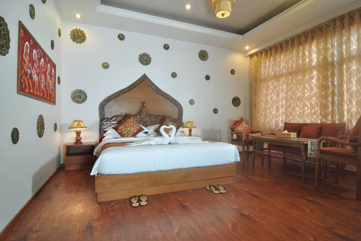A well-furnished bedroom with a large bed, a wooden headboard, decorative wall art, a seating area with a table and chairs, and a window with curtains.