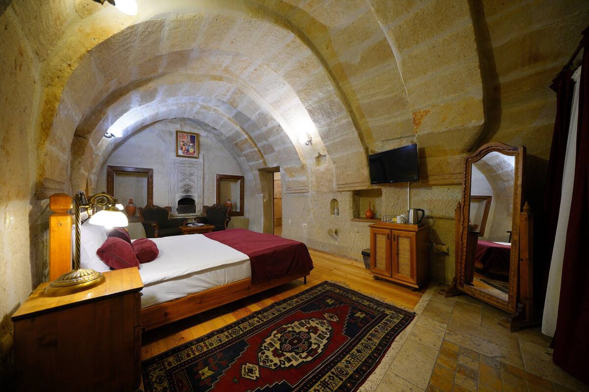 Ein Zimmer mit gewölbter Decke, einem Bett mit roter Bettdecke, einem Nachttisch aus Holz, einer Lampe, einem an der Wand montierten Fernseher und einem Spiegel.