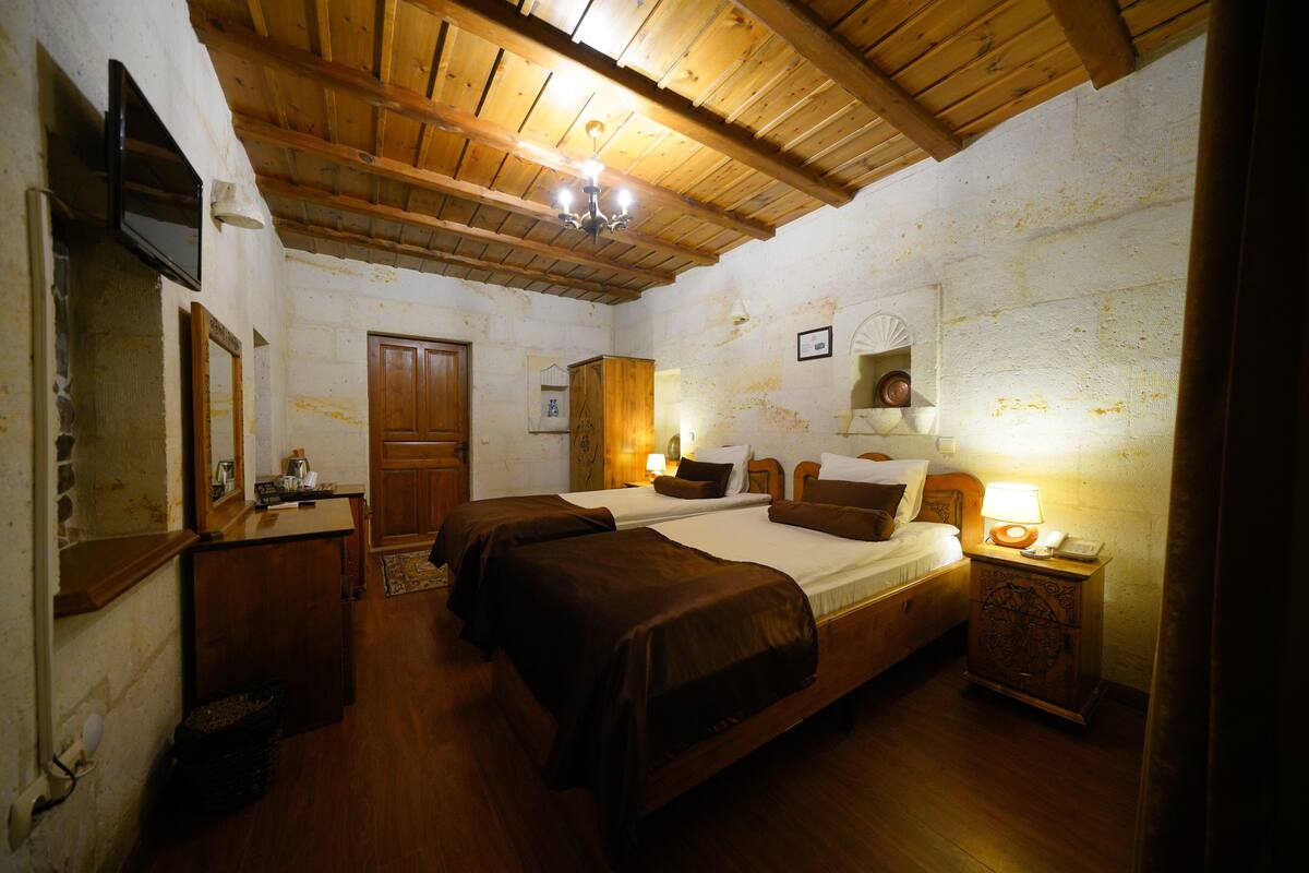 Ein Zimmer mit zwei Betten, Holzbalkendecke, einem Holzschrank, einem Nachttisch mit Lampe und einem an der Wand montierten Fernseher.