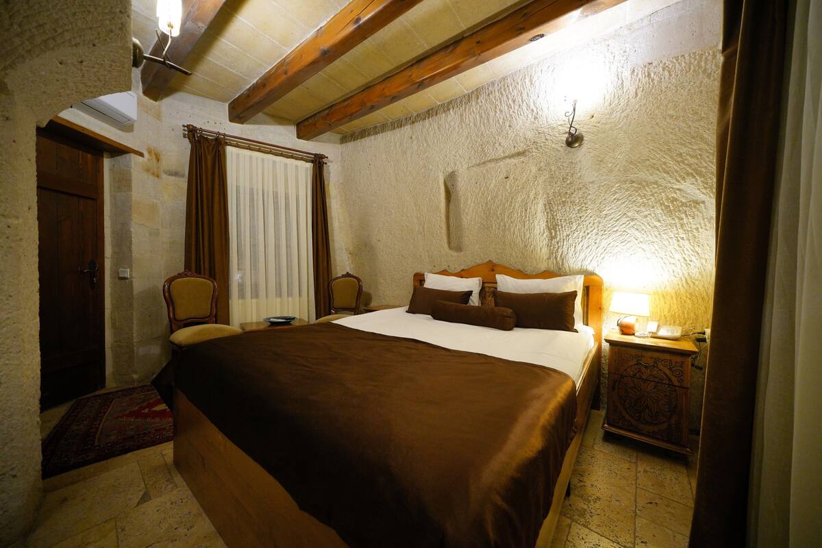 Ein Zimmer mit einem großen Bett, einem Holzkopfteil, einem Nachttisch mit Lampe, einem Sessel und einem Fenster mit Vorhängen.