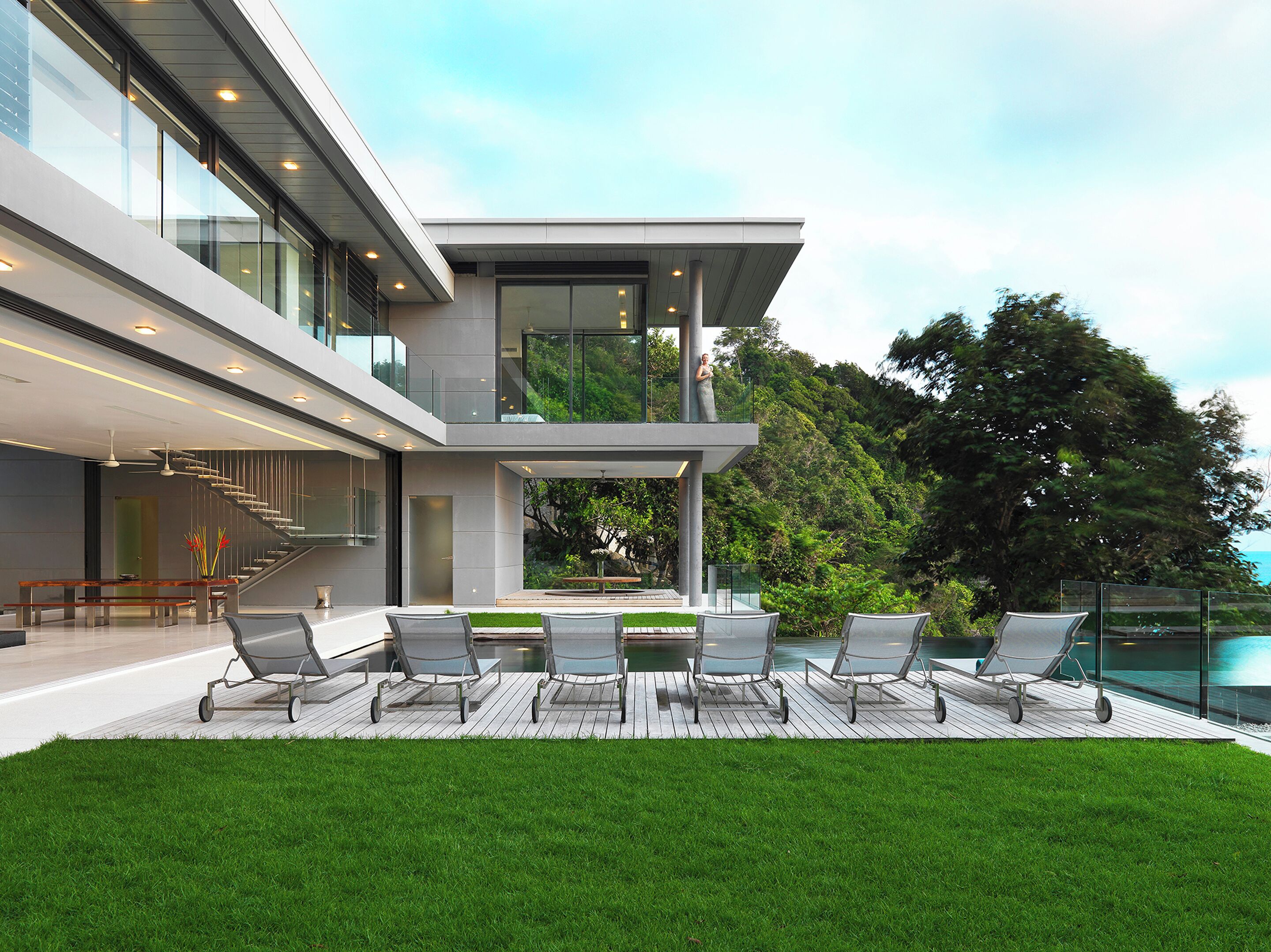 A modern two-story house with a balcony, outdoor seating area, and a view of the surrounding greenery.