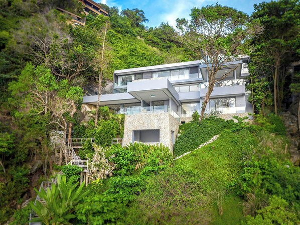 A modern multi-story house with a stone facade, surrounded by lush greenery and trees.