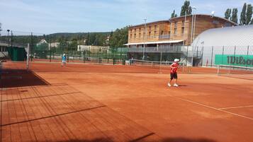 A tennis court with a net, surrounded by a fence. There are buildings in the background.