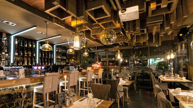 Ein modernes Restaurant-Interieur mit Barhocker-Bestuhlung, hohen Decken und dekorativer Beleuchtung.