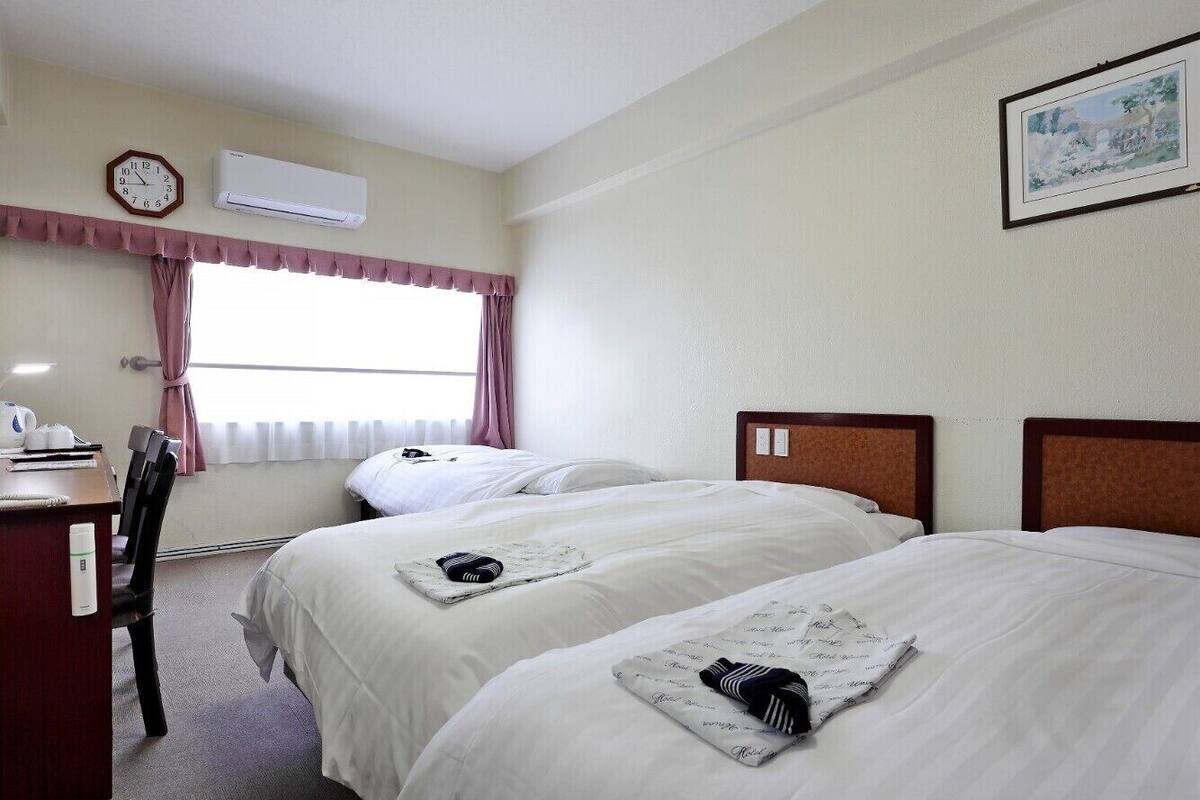 Une chambre d’hôtel avec deux lits, un bureau, une chaise, une horloge, un climatiseur et une fenêtre avec des rideaux.