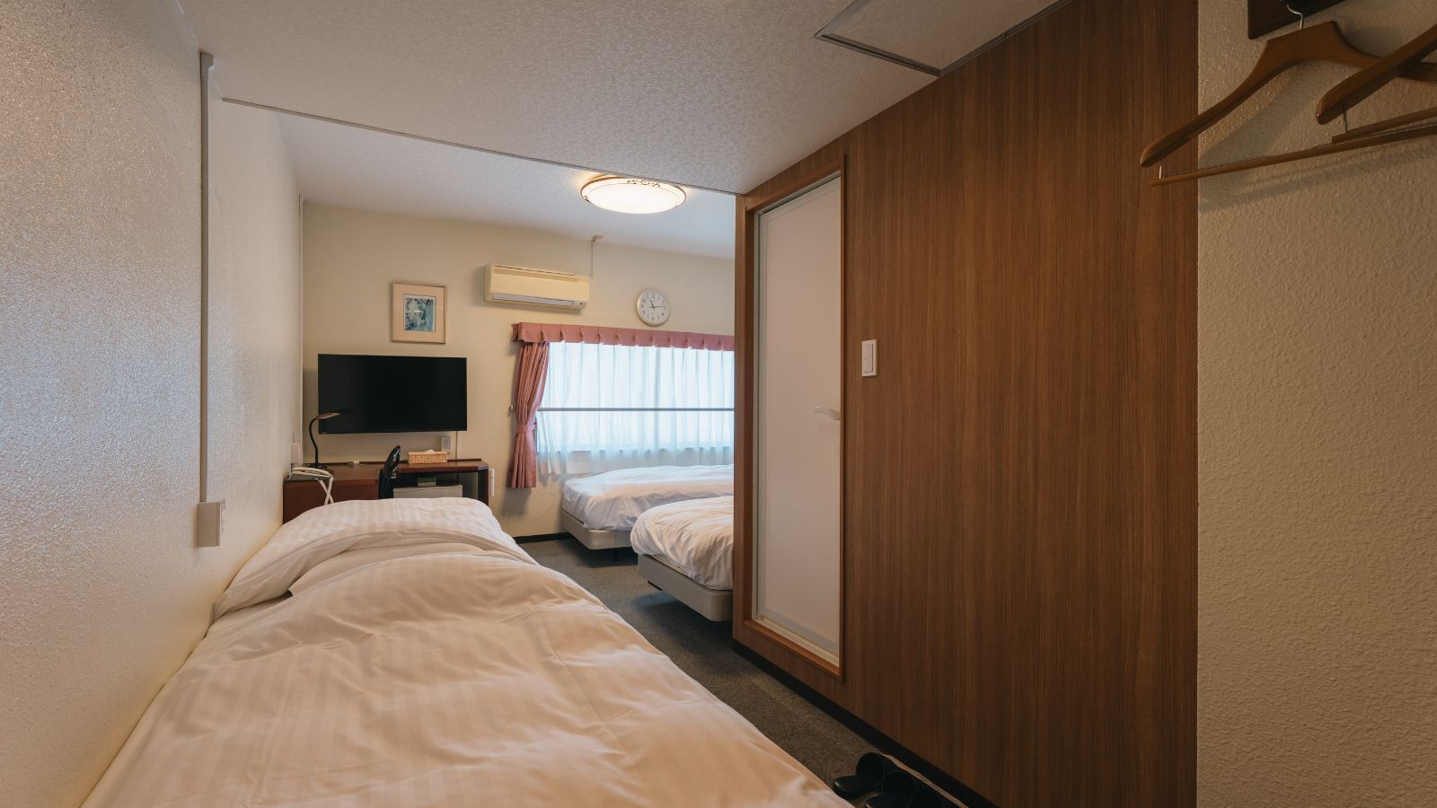 Een hotelkamer met twee bedden, een televisie, een airconditioner, een klok en een houten garderobe.