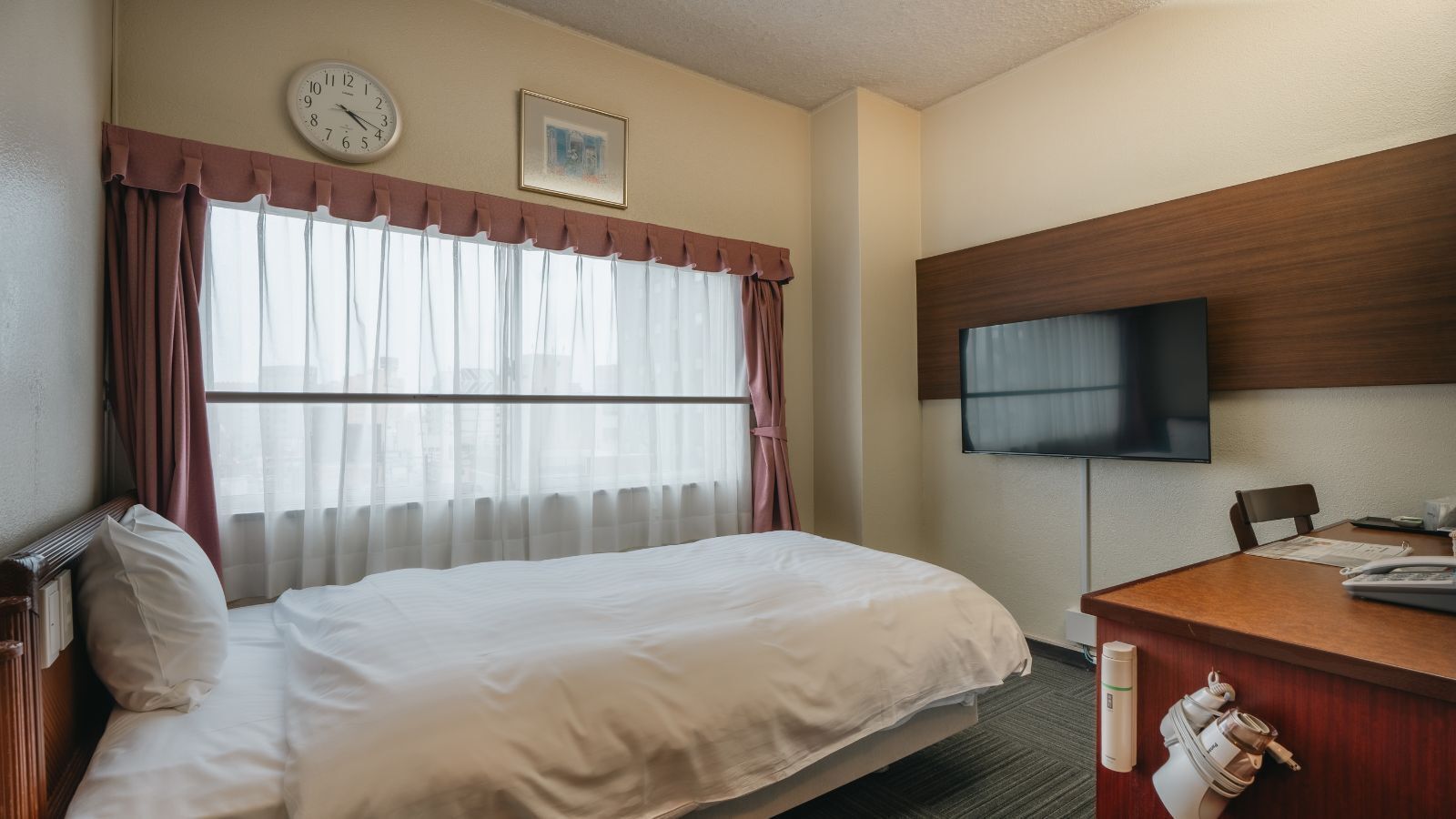 Een hotelkamer met een bed, een bureau met telefoon, een televisie, een klok en een raam met gordijnen.
