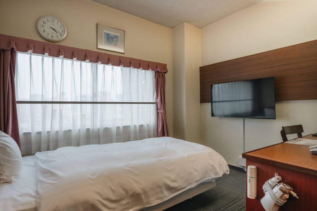 Une chambre d’hôtel avec un lit, un bureau avec un téléphone, une télévision, une horloge et une fenêtre avec des rideaux.