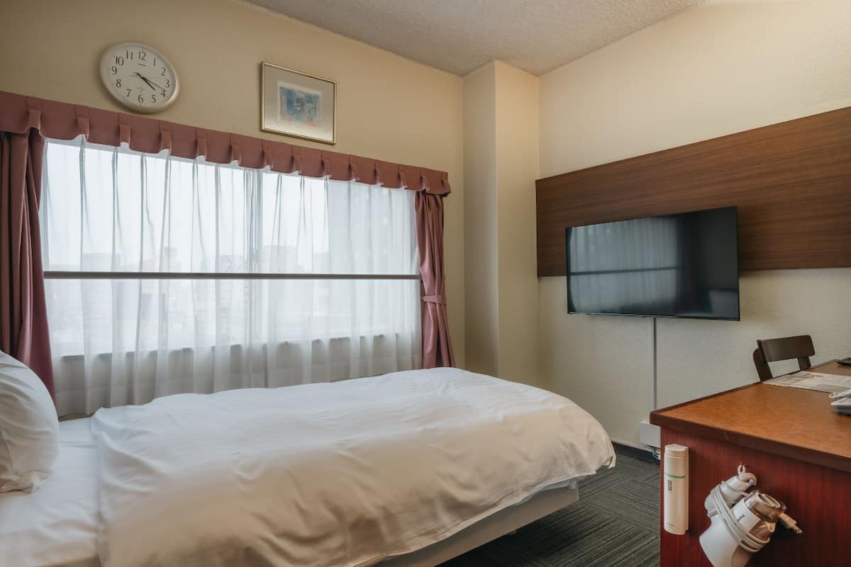 A hotel room with a bed, a desk with a telephone, a television, a clock, and a window with curtains.