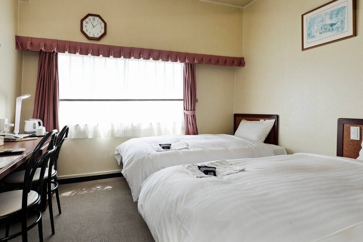Une chambre d’hôtel avec deux lits, un bureau avec des chaises, une horloge et une fenêtre avec des rideaux.
