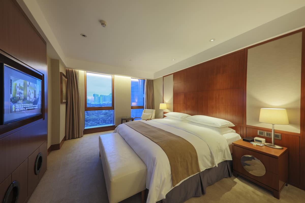 A hotel room with a large bed, a bedside table with a lamp, a TV mounted on the wall, and a window with curtains.