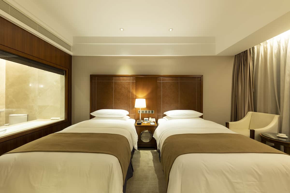 A hotel room with two beds, a large window, a round clock, and a lamp on the nightstand.