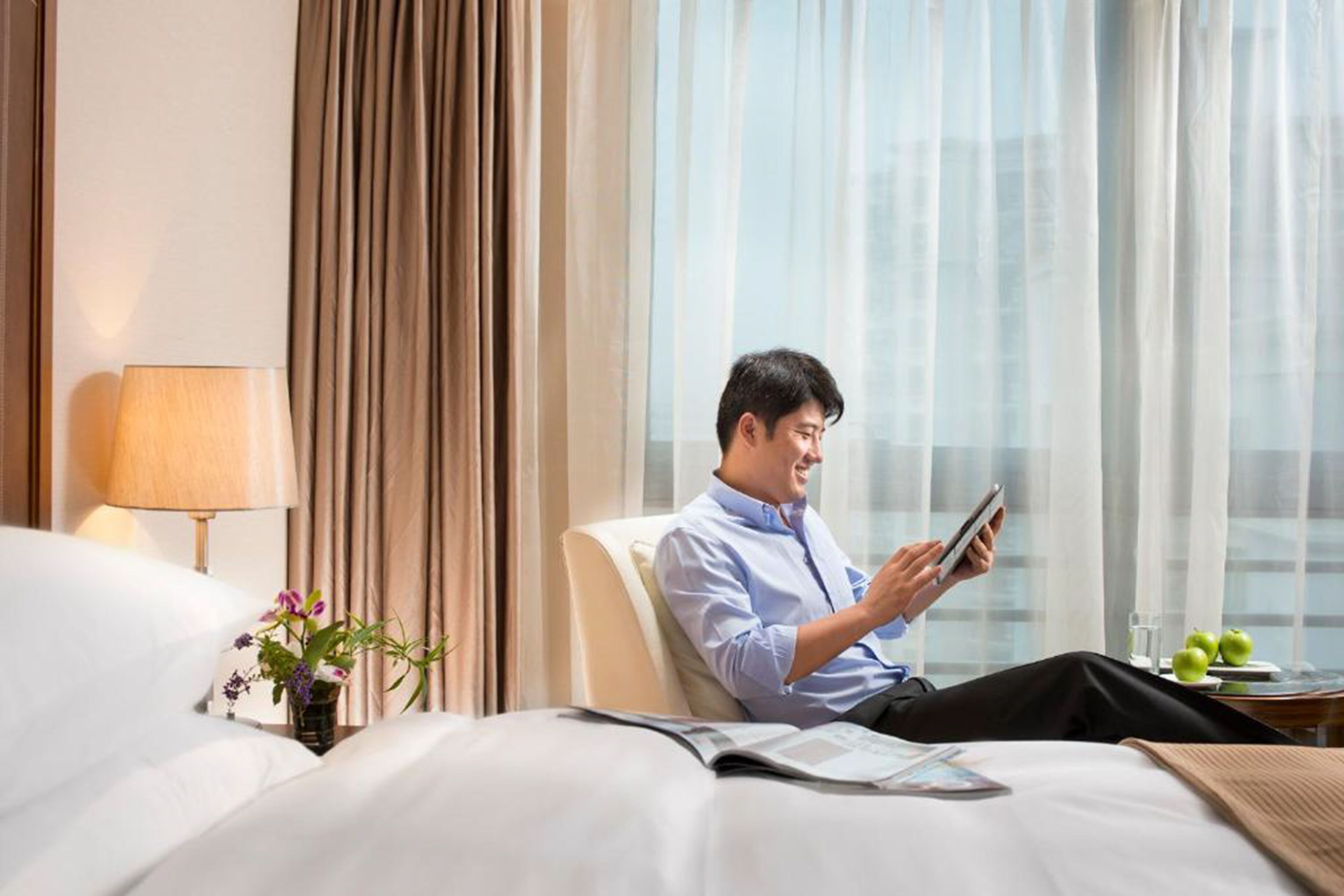 A man sitting on a bed in a hotel room, reading a book.