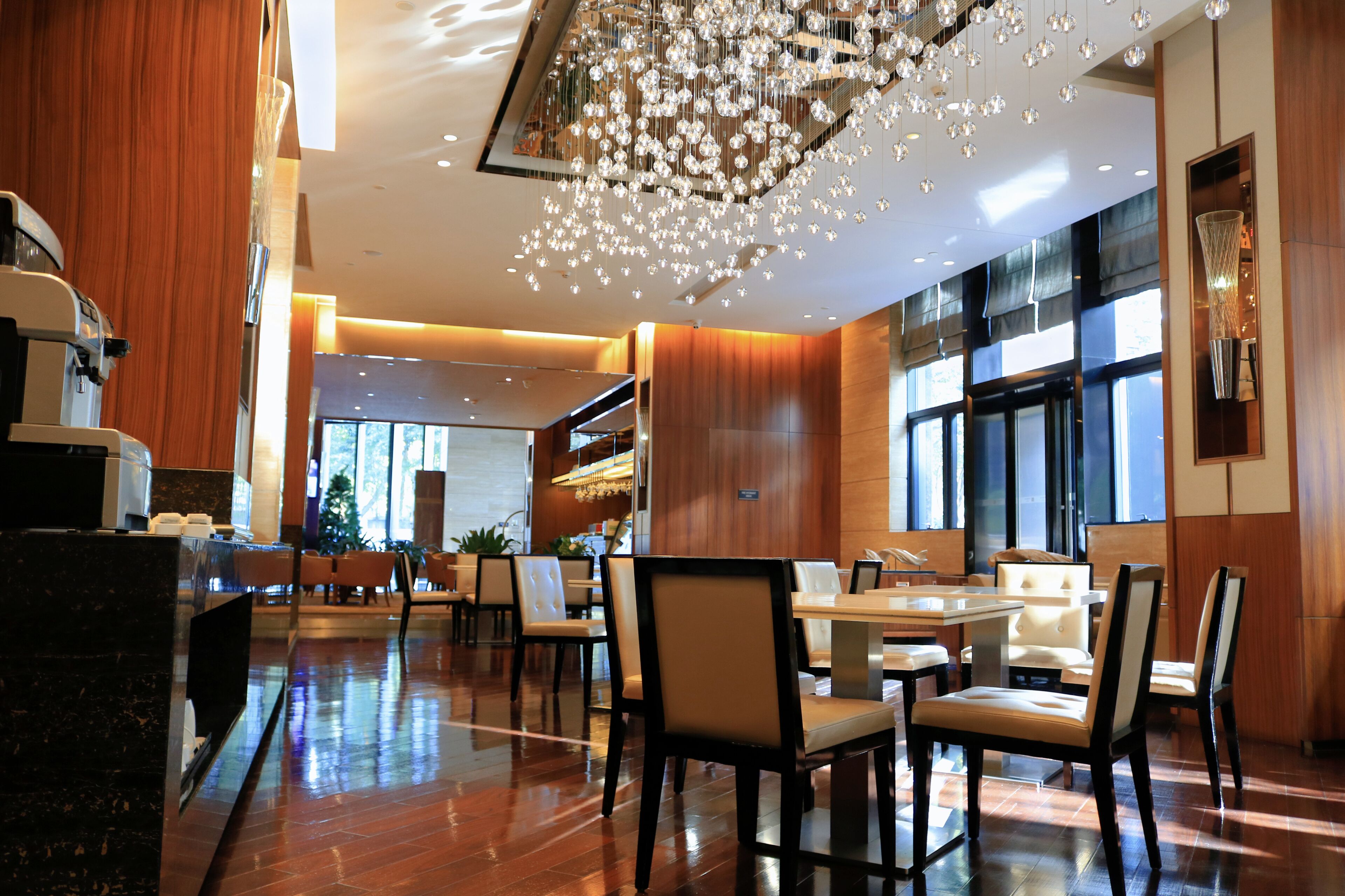 A modern hotel lobby with a wooden reception desk, seating area with tables and chairs, and a large chandelier.