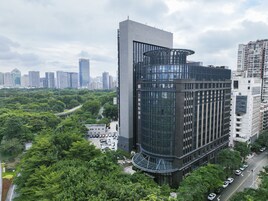 A modern hotel with a curved glass facade, surrounded by lush greenery and adjacent to a busy road.