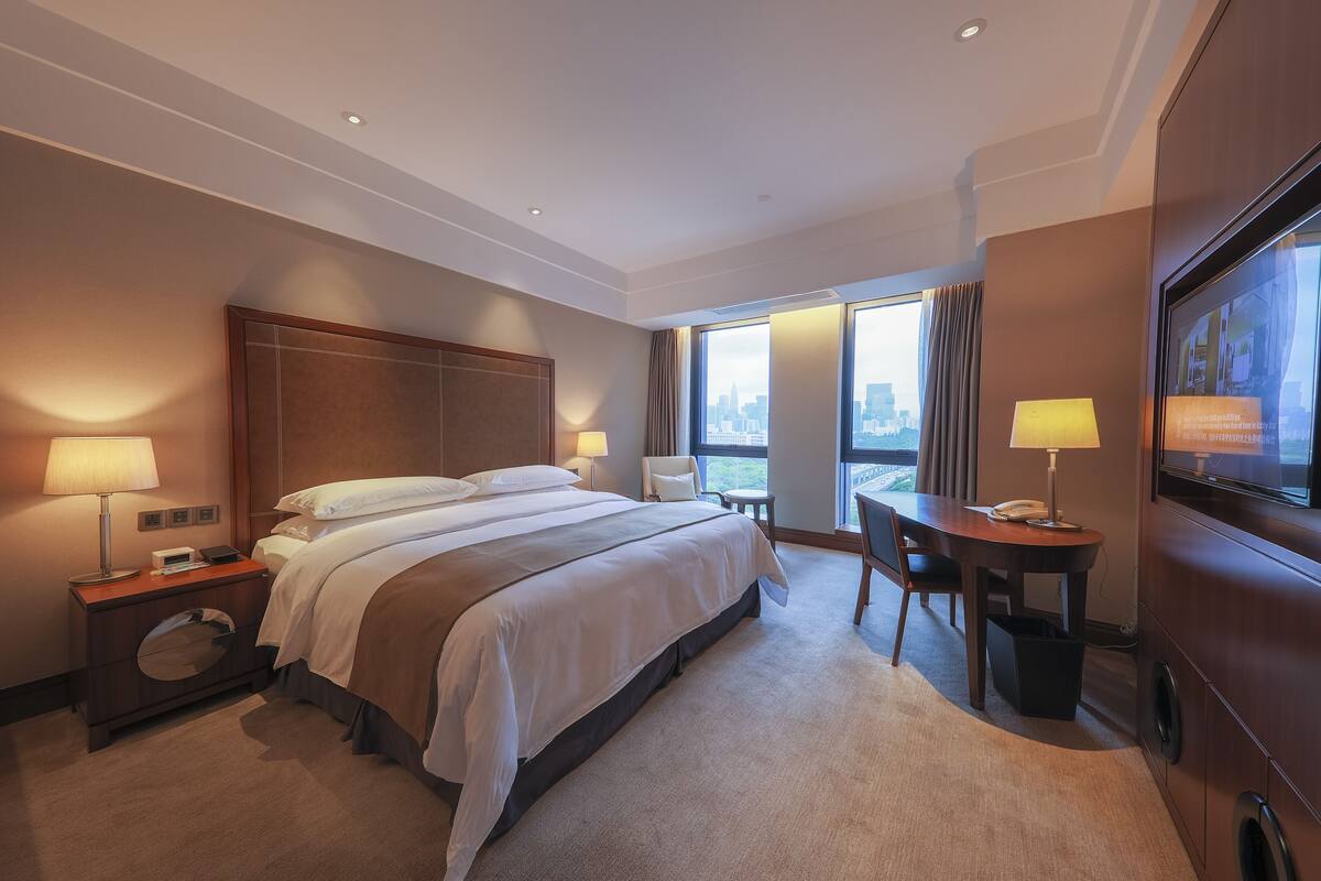 A hotel room with a large bed, a desk, a chair, a TV, and a view of the city through the windows.