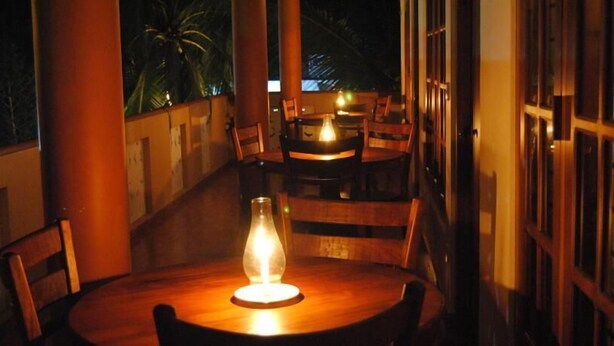 A dimly lit outdoor seating area with wooden tables and chairs, a glass bottle candle, and a view of palm trees. - Kumara Guesthouse Unawatuna (Unawatuna)
