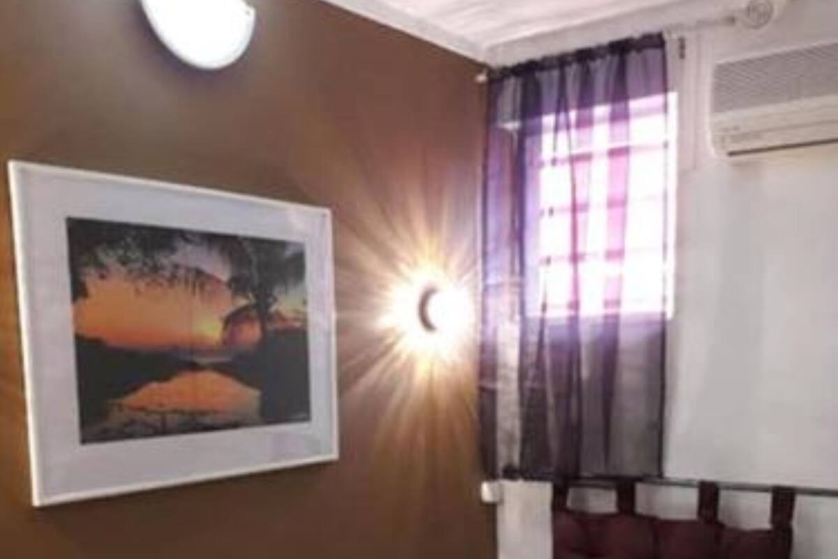 A single bed with a headboard, a wall-mounted air conditioning unit, a window with curtains, and a framed picture on the wall.